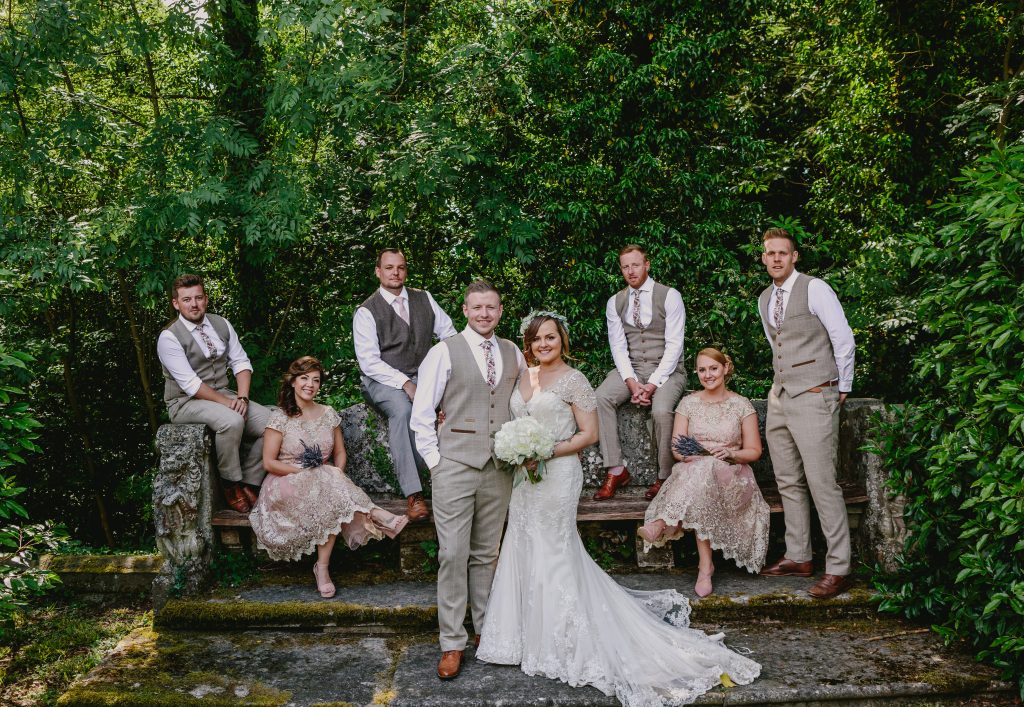 A Bridal party of 8, sit on a bench in the grounds of Hampton Manor