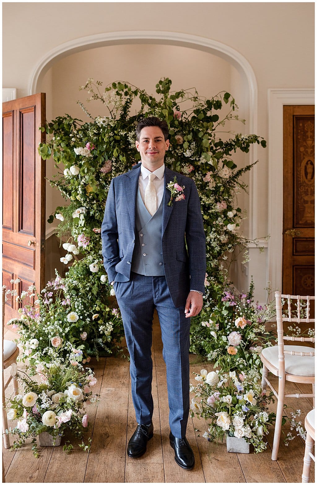 Groom stands in front of a floral arch before his Alrewas Hayes wedding ceremony