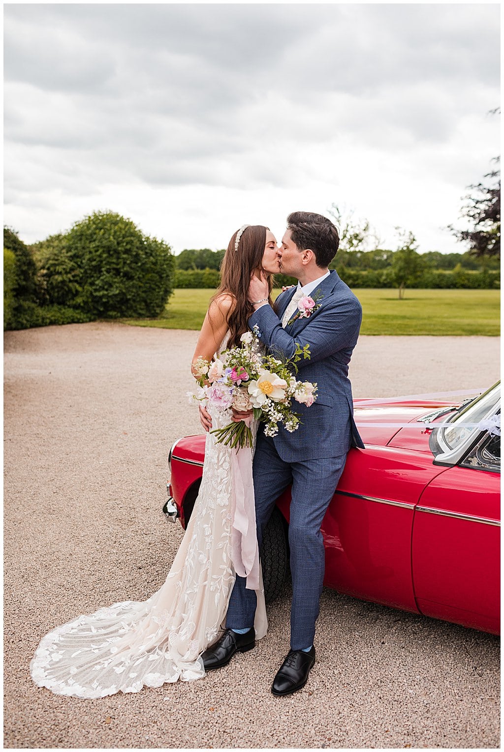 Bride wearing strappy lace wedding dress kissing her Groom, sitting on a red sports car 