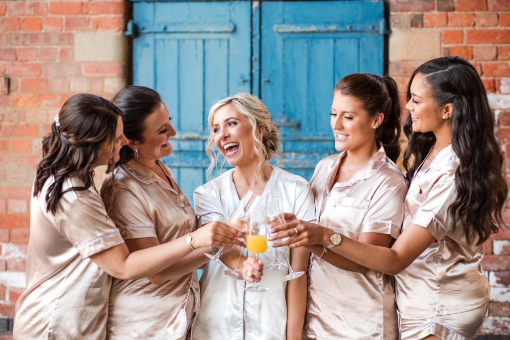 Bride and Bridesmaids wearing matching pyjamas have a toast in front of a blue door at a Thorpe Garden wedding