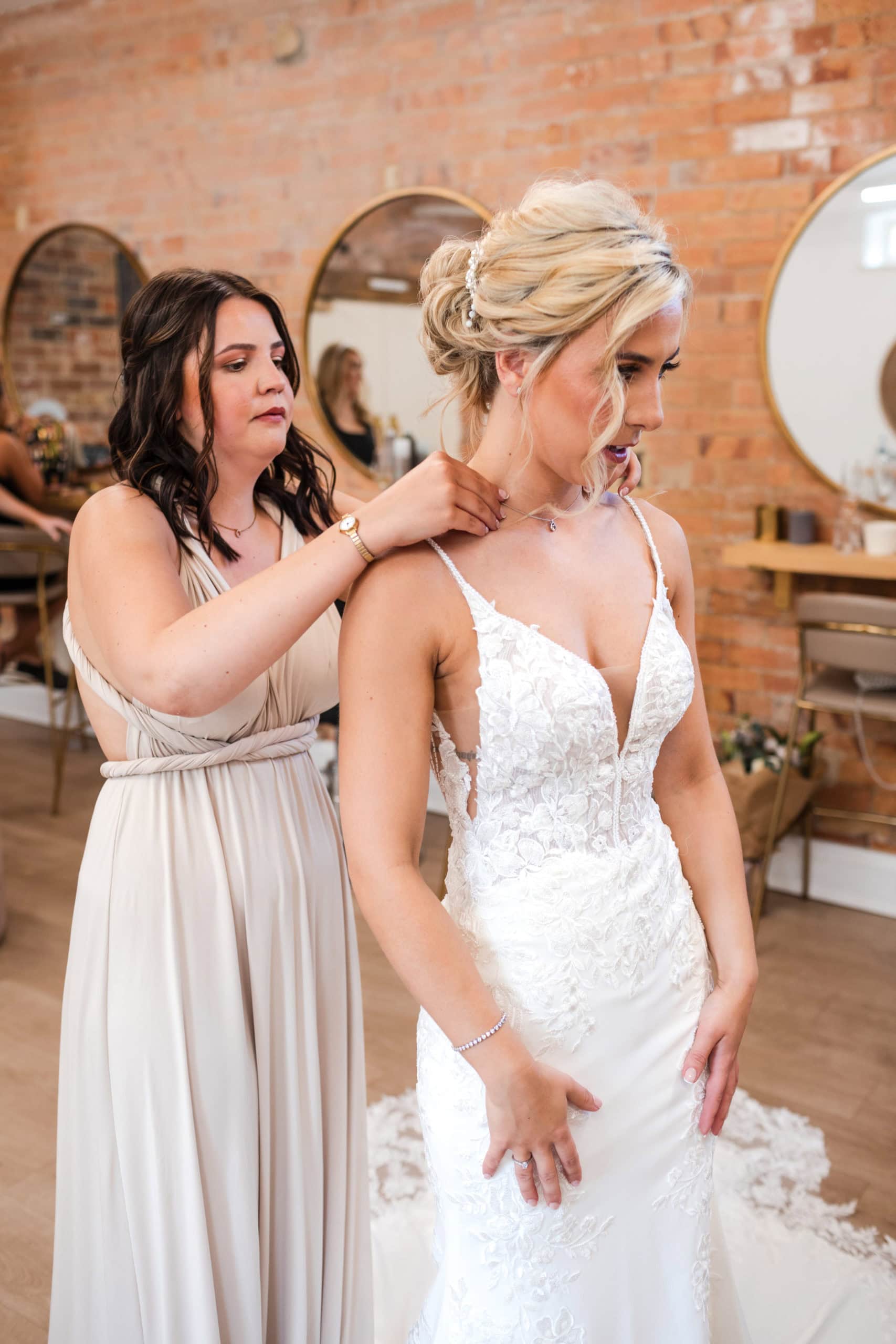 Bridesmaid waring nude coloured wrap dress, helps the bride put on her necklace, getting ready at a Thorpe Garden wedding