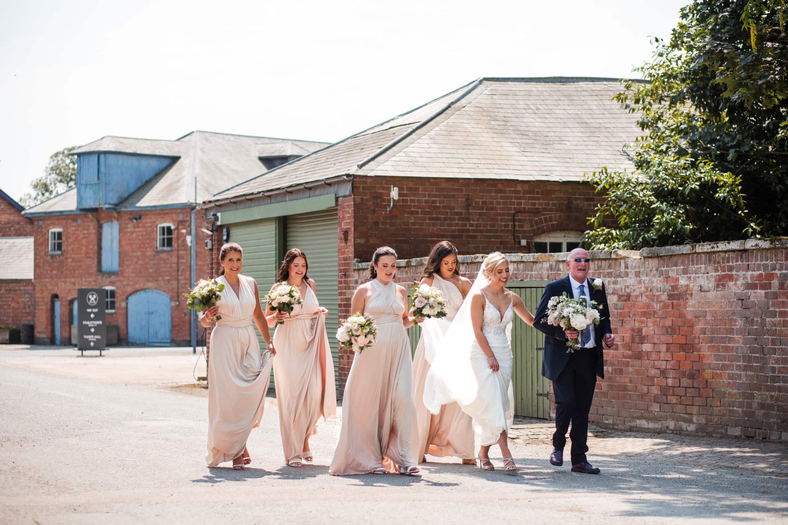 A Bride holds her Dads arm and walks with her Bridesmaids to her wedding at Thorpe Garden. Bridesmaids wear nude coloured wrap dresses