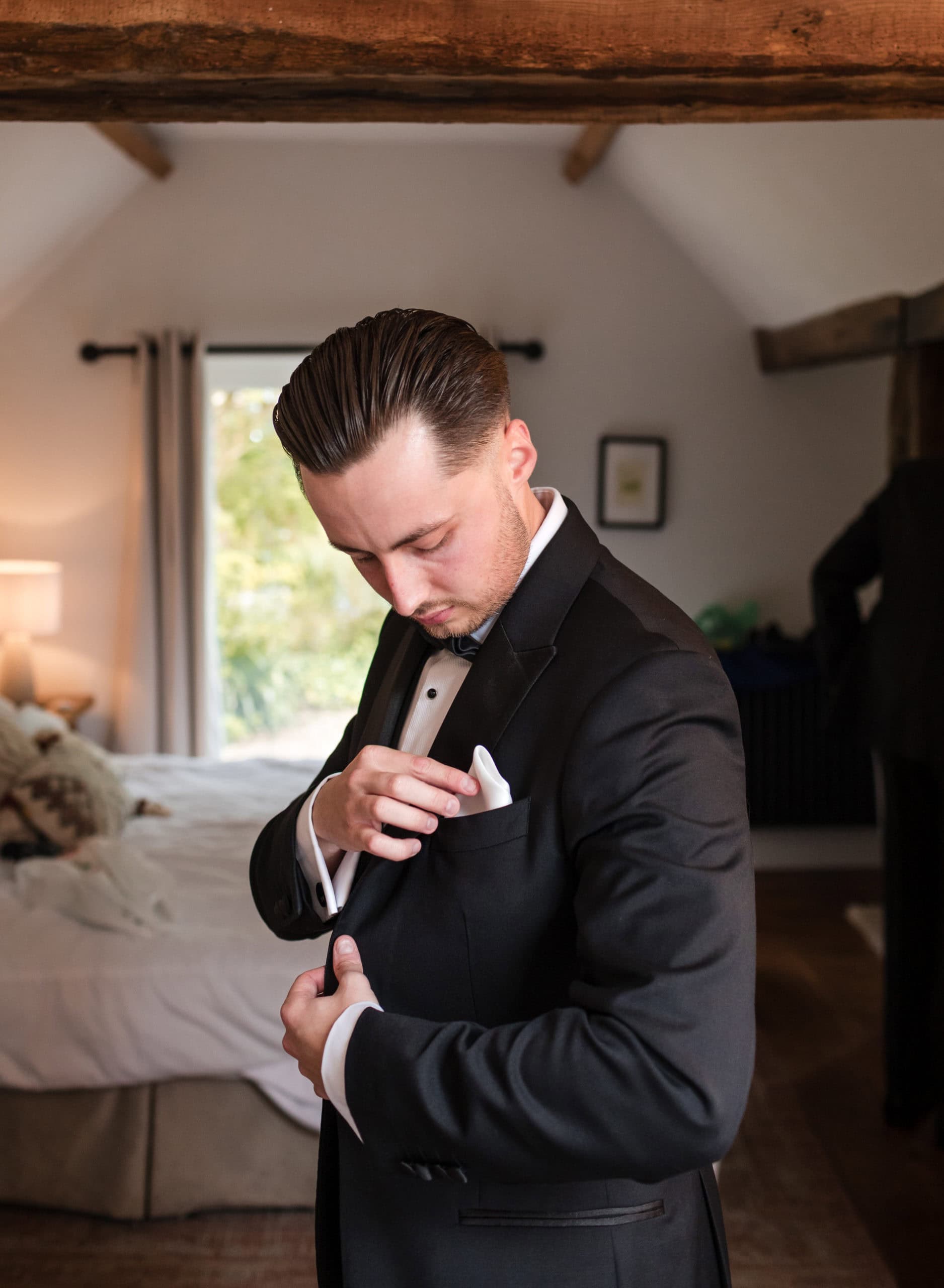 Groom wearing a tuxedo makes adjustment to his pocket square in the honeymoon cottage at Curradine Barn