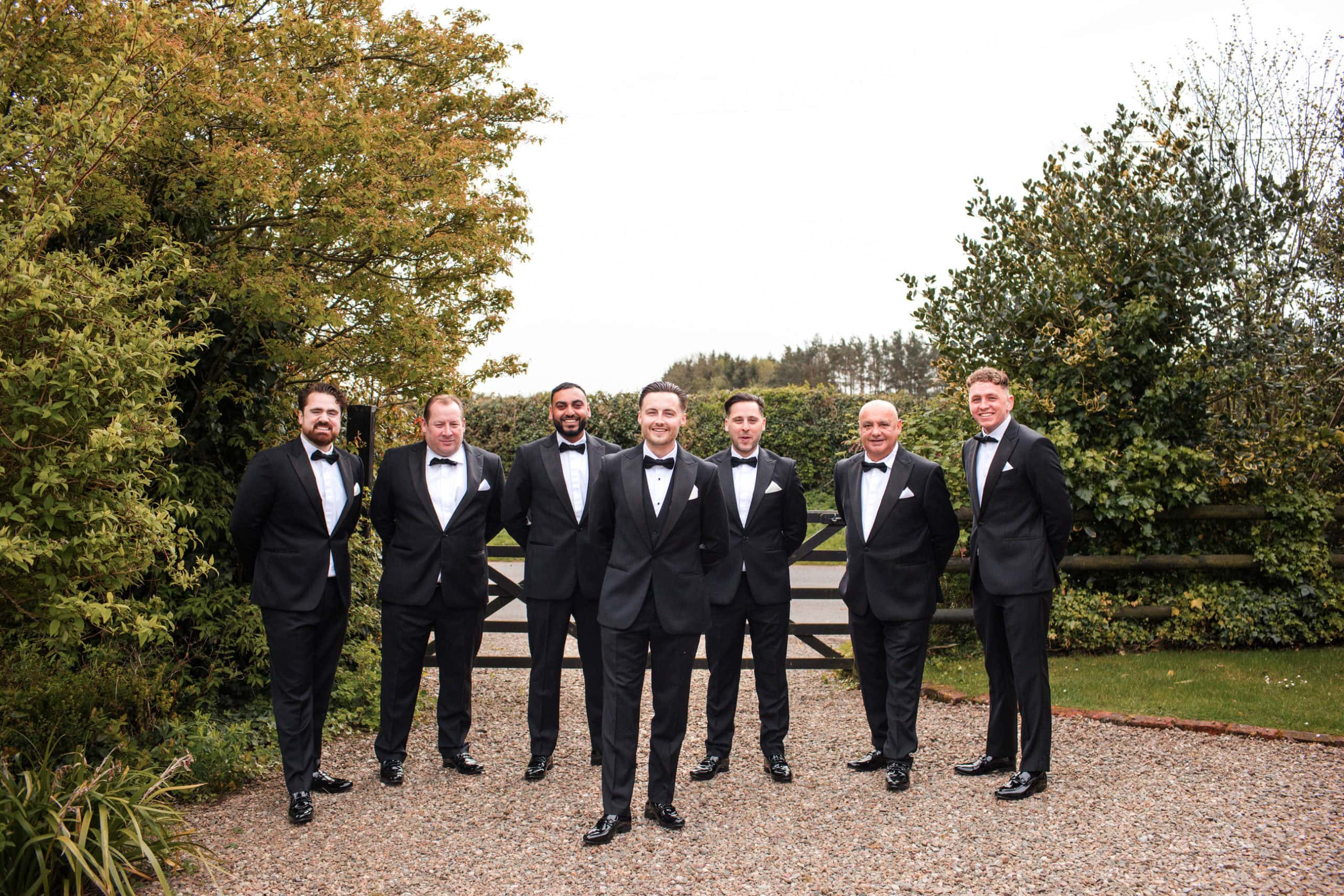 Groom and six groomsmen standing in front of the farmhouse gate at Curradine Barn, wearing black tie