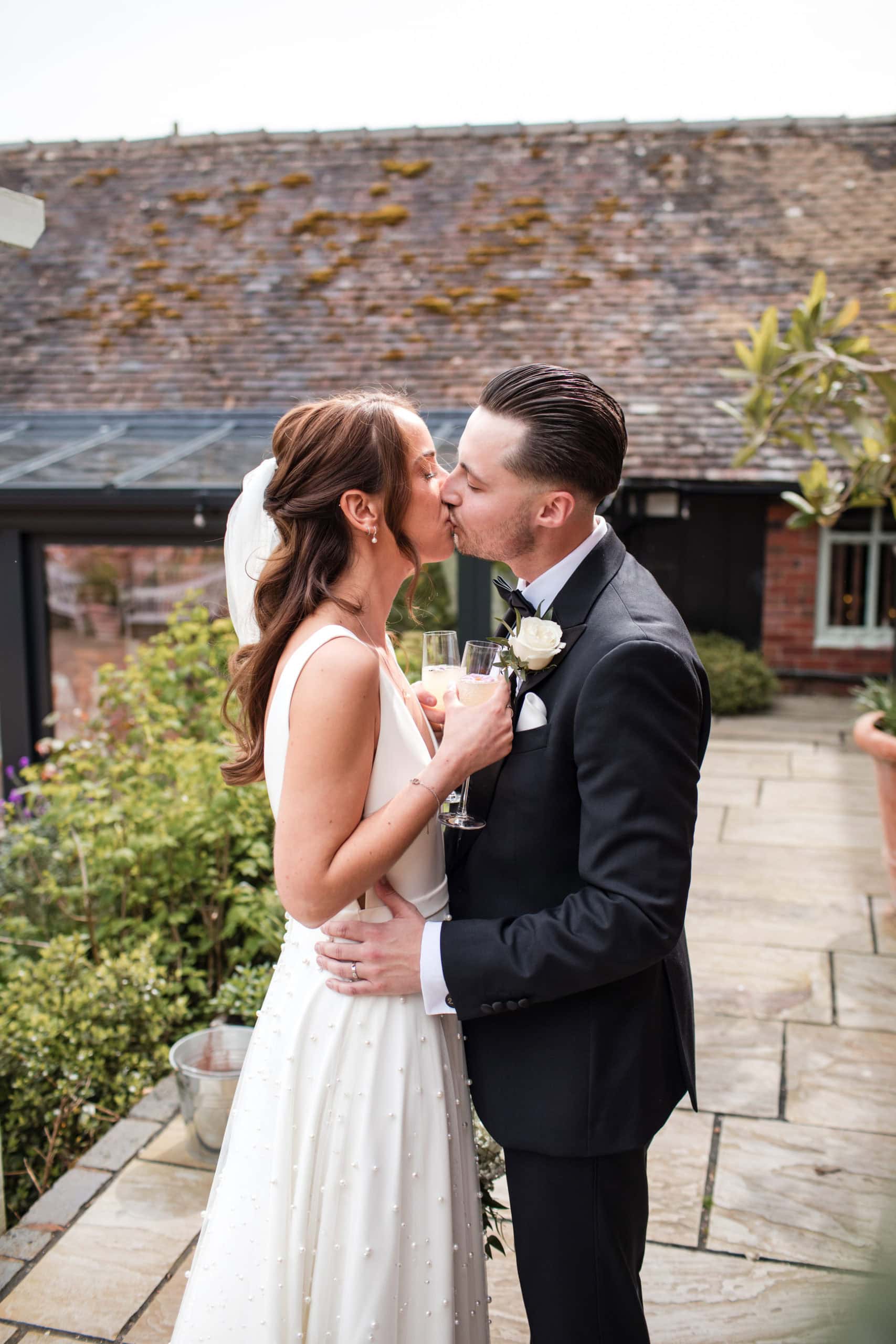 Couple stand in the courtyard at Curradine Barns and kiss holding drinks after their wedding ceremony