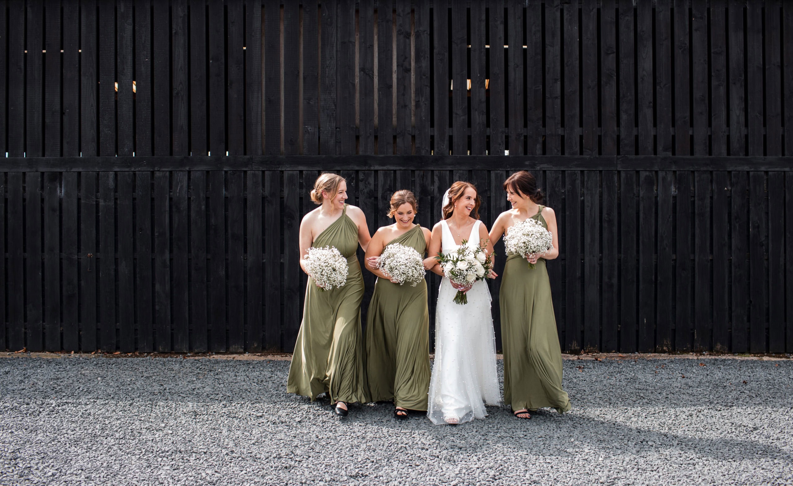 Bride and her bridesmaids link arms and walk in front of a black wooden barn laughing