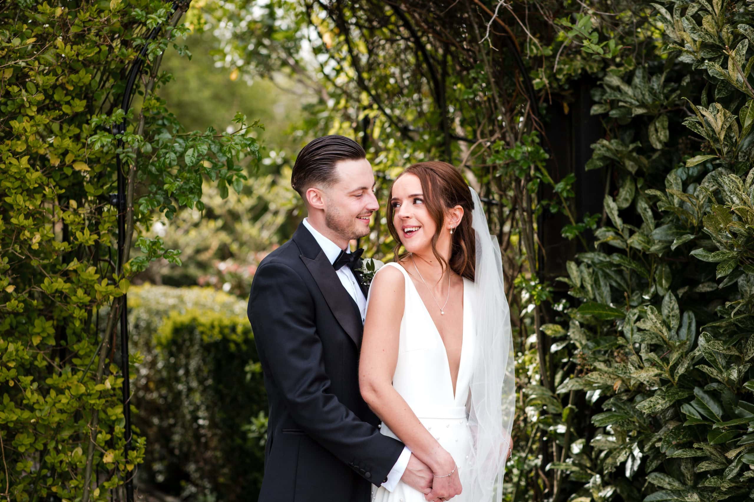 Groom stands behind Bride holding hands under an arch of trees. Bride looking back at Groom