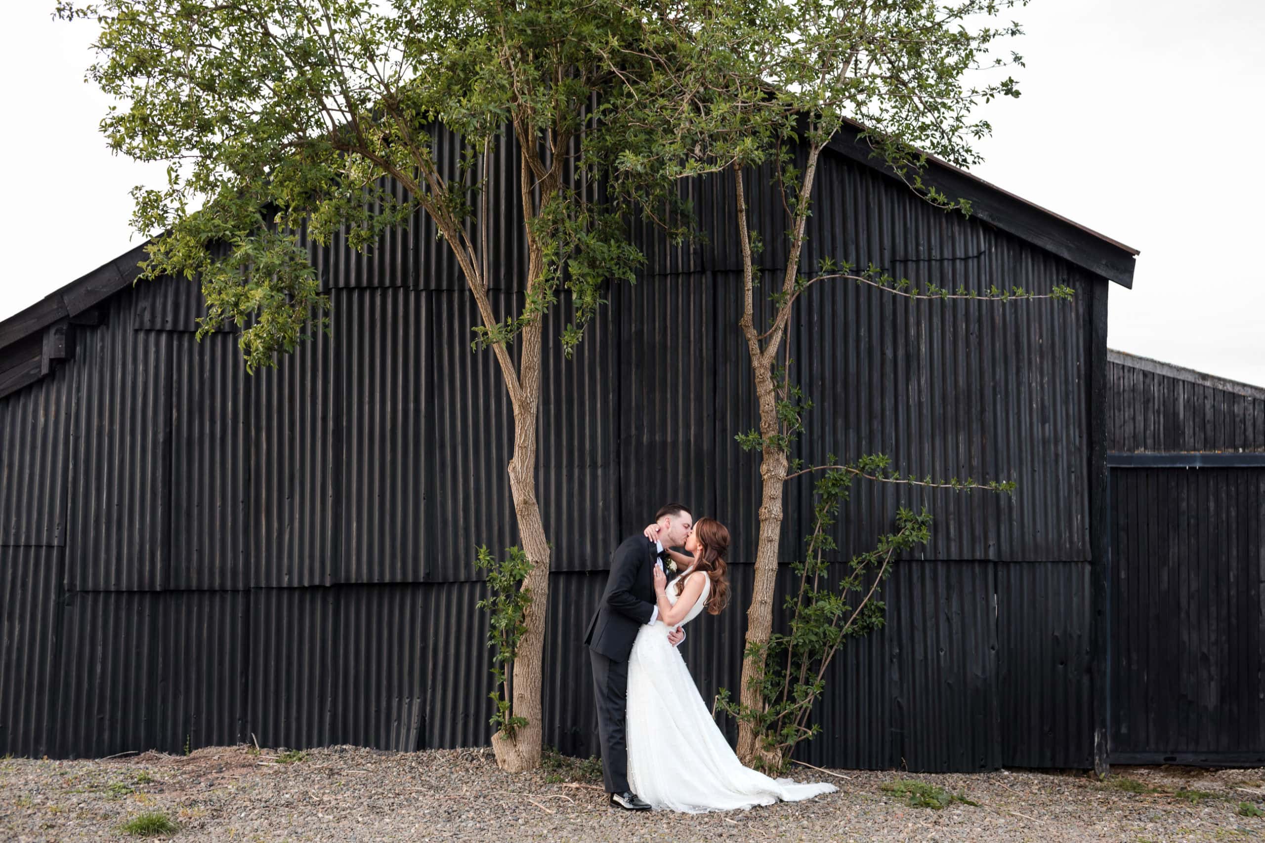 Couple dip and kiss standing in front of a black corrugated iron barn in Worcestershire countryside