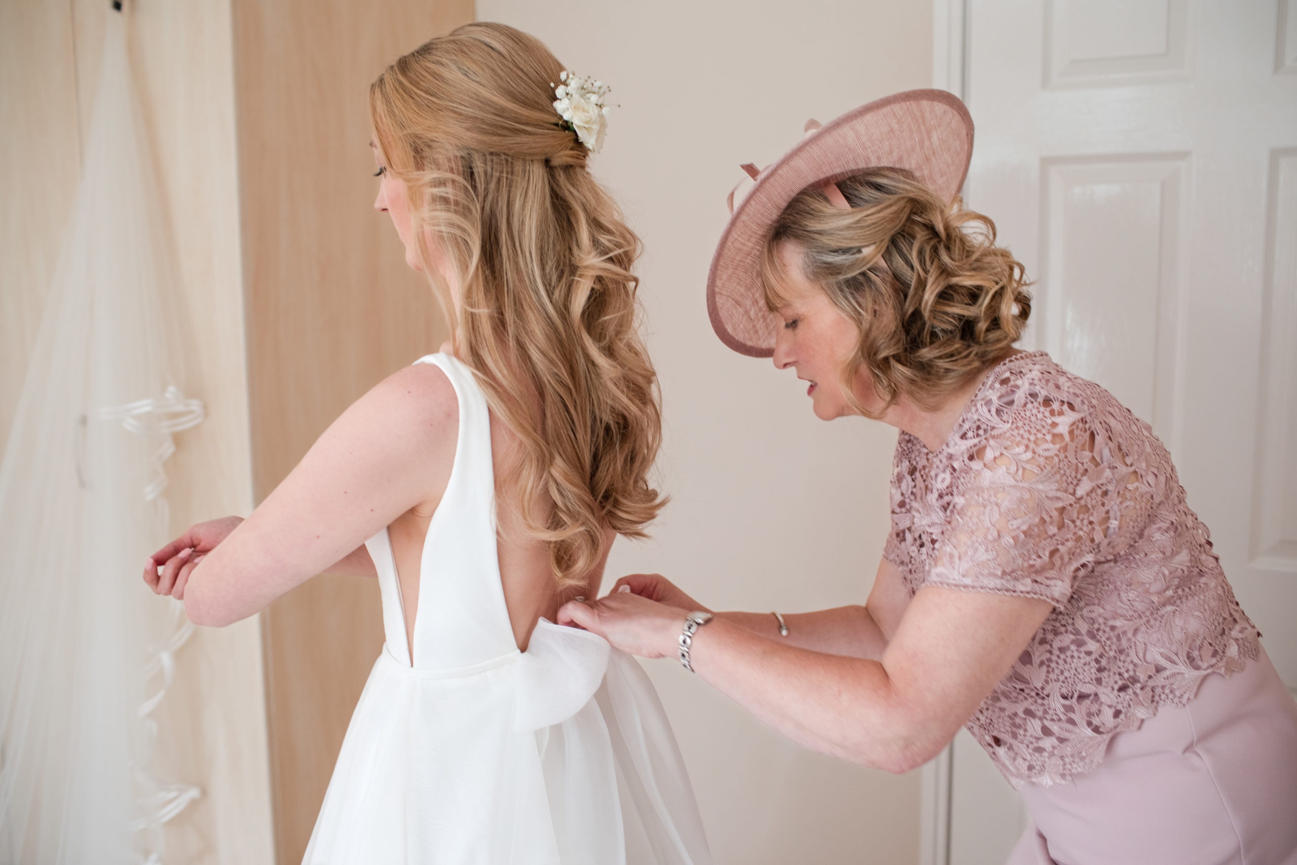 Mother of the Bride wearing pink hat buttons the back of her Daughters wedding dress