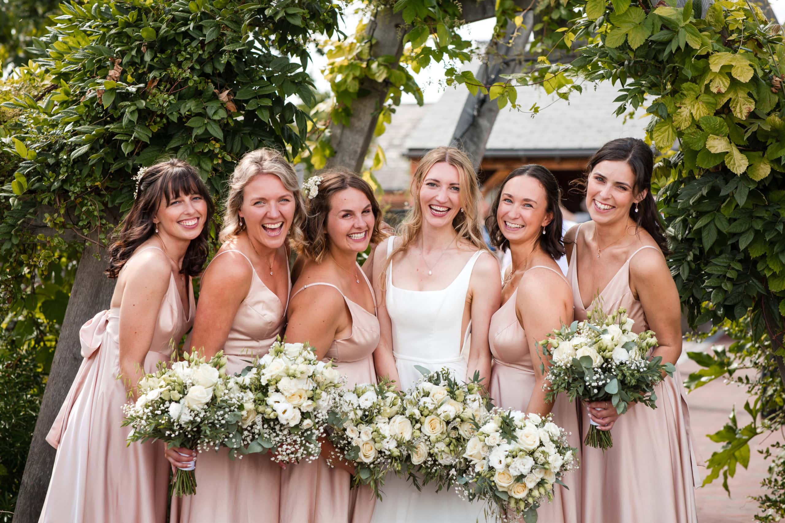 Bride and five Bridesmaids wearing blush pink stripy dresses, stand infant of foliage covered archway at Cider Mill Barn wedding