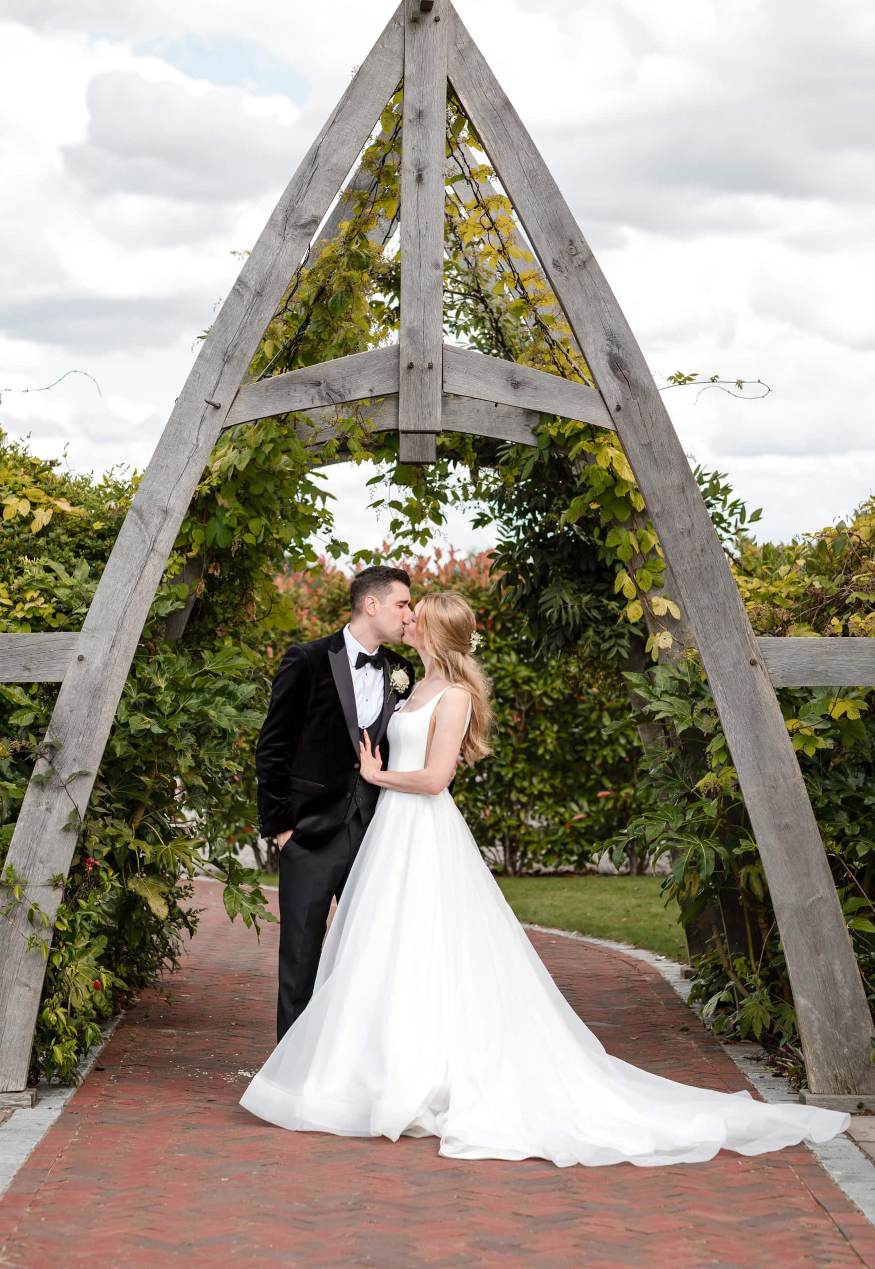 Bride & Groom stand under a foliage covered wooden archway and share a kiss. Groom wears a tuxedo