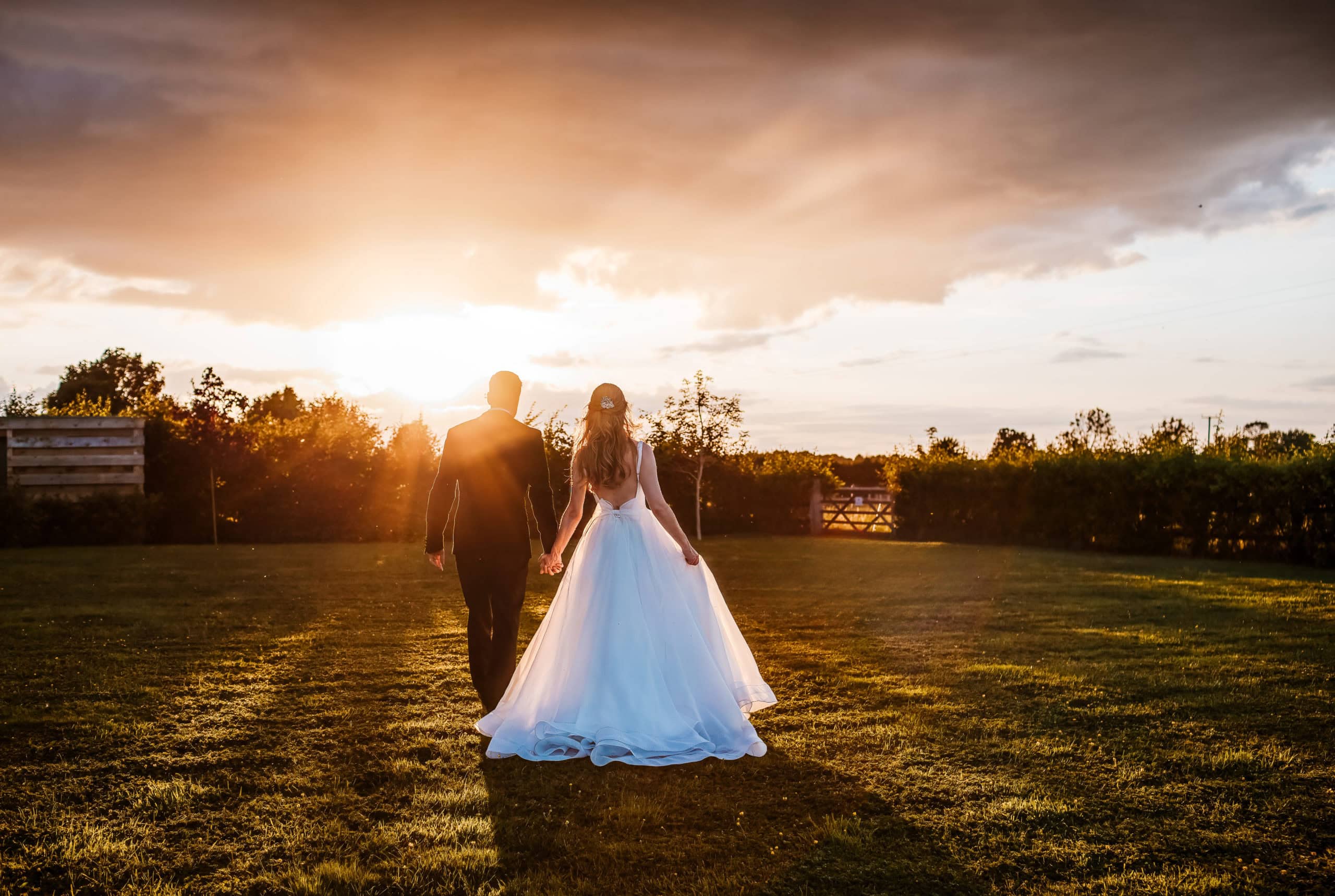 Bride and Groom hold hands and walk across a field into the sunset