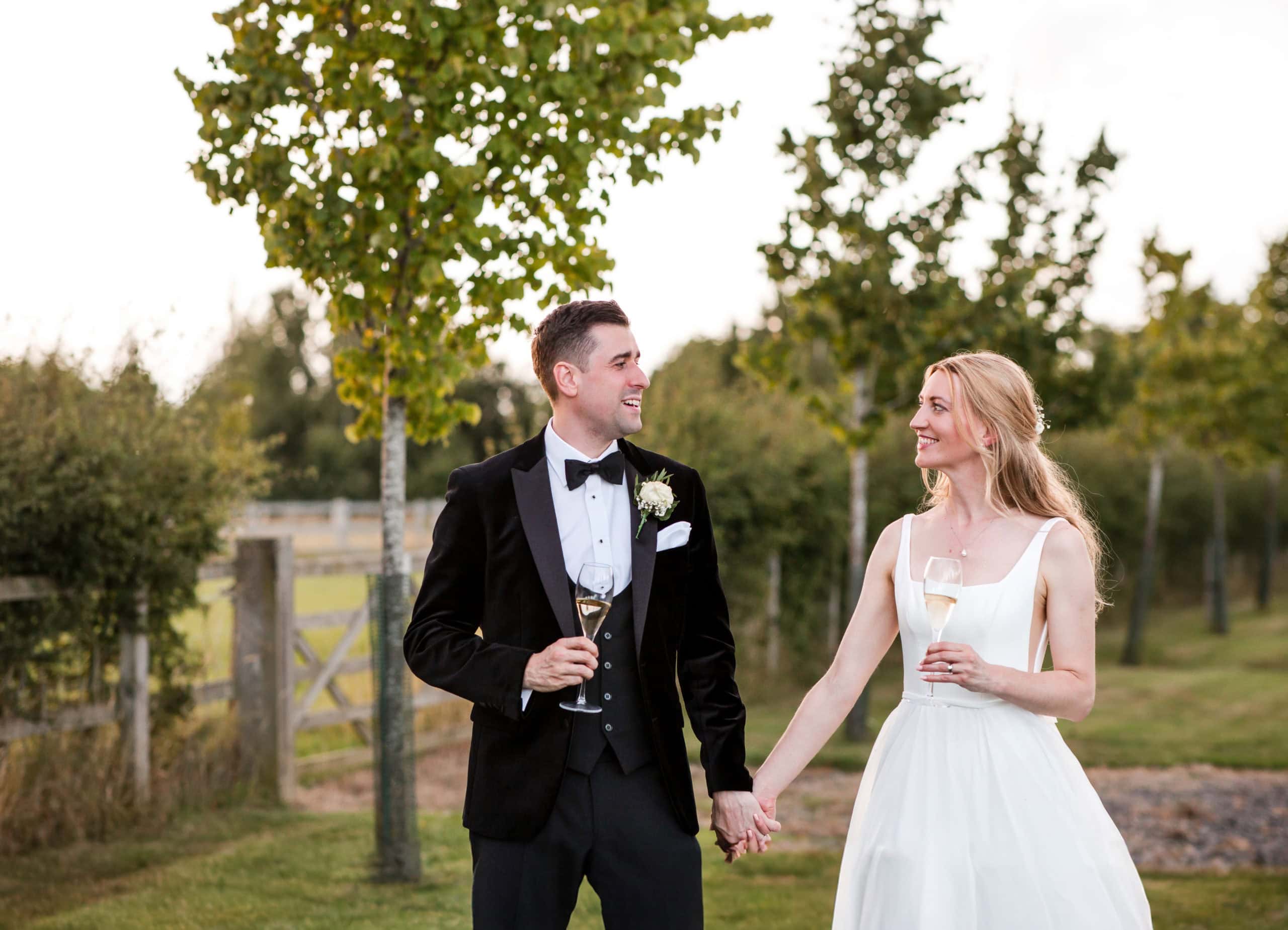 Just married couple hold hands and walk with glasses of champagne. Groom wears a black velvet tuxedo and bow tie