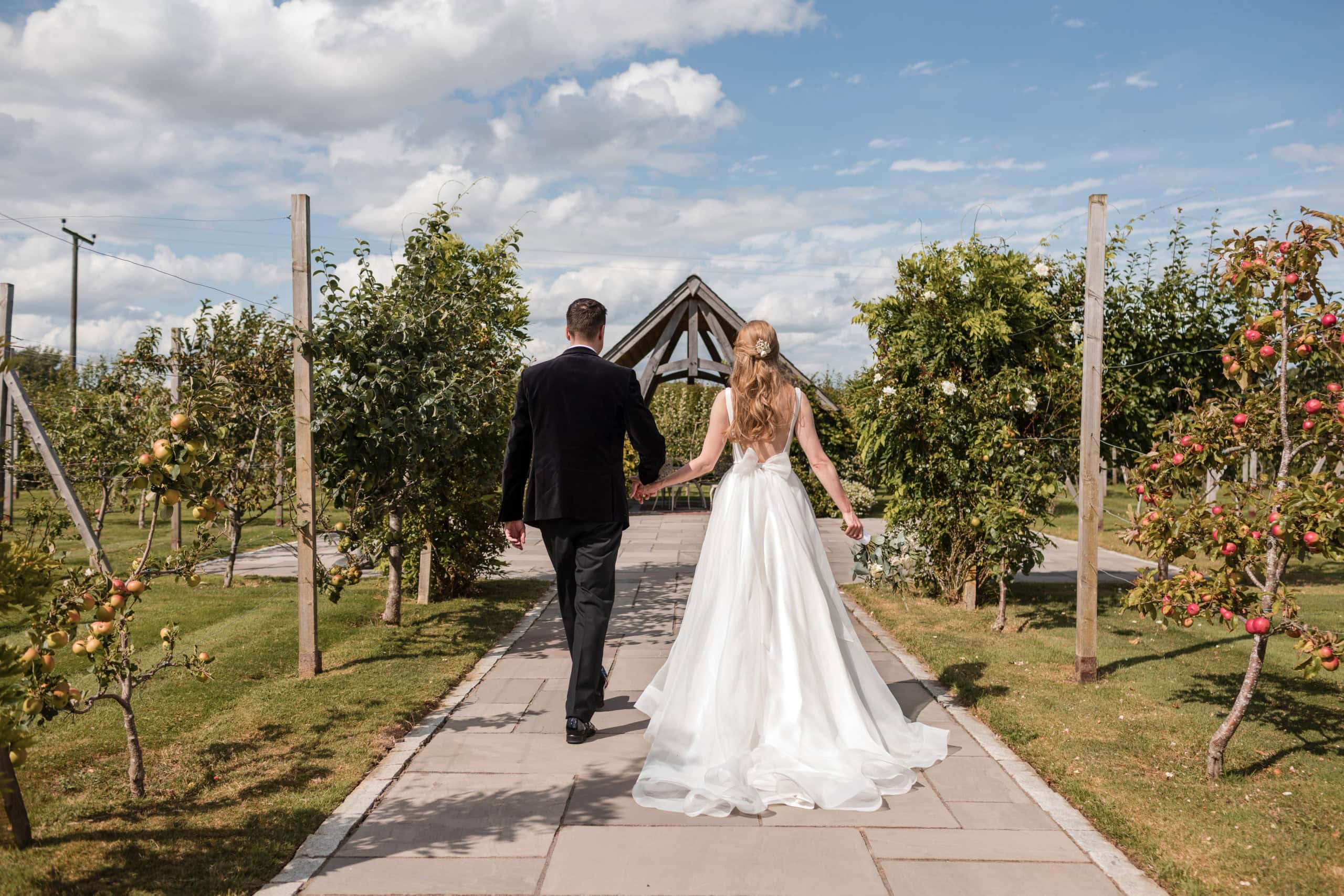 couple hold hands and walk towards the summer house in the orchard at cider mill barn