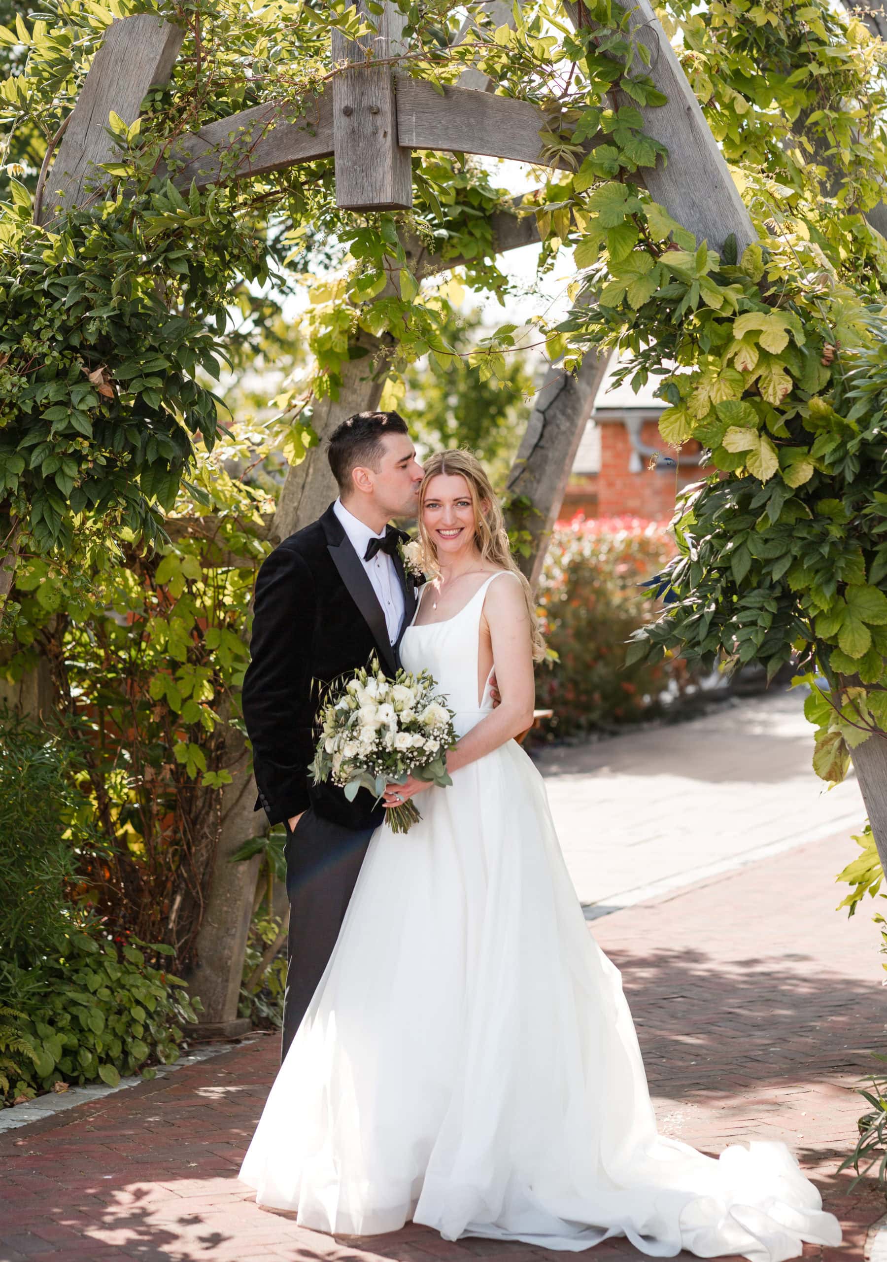 A groom wearing black tie kisses his Bride on the forehead. Bride holds a white and green bouquet