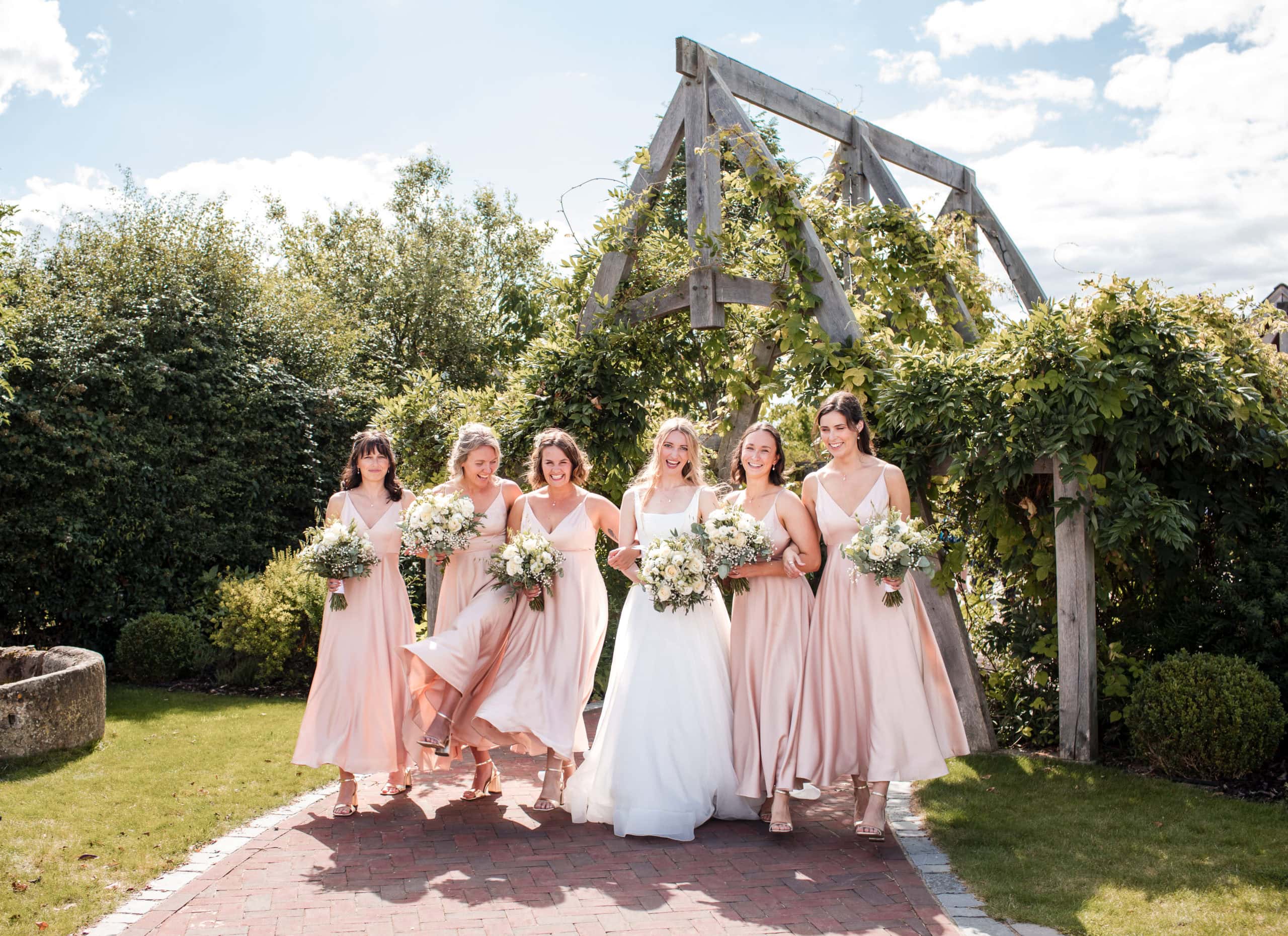 Bride and five bridesmaids wearing blush pink, link arms to walk and laugh together