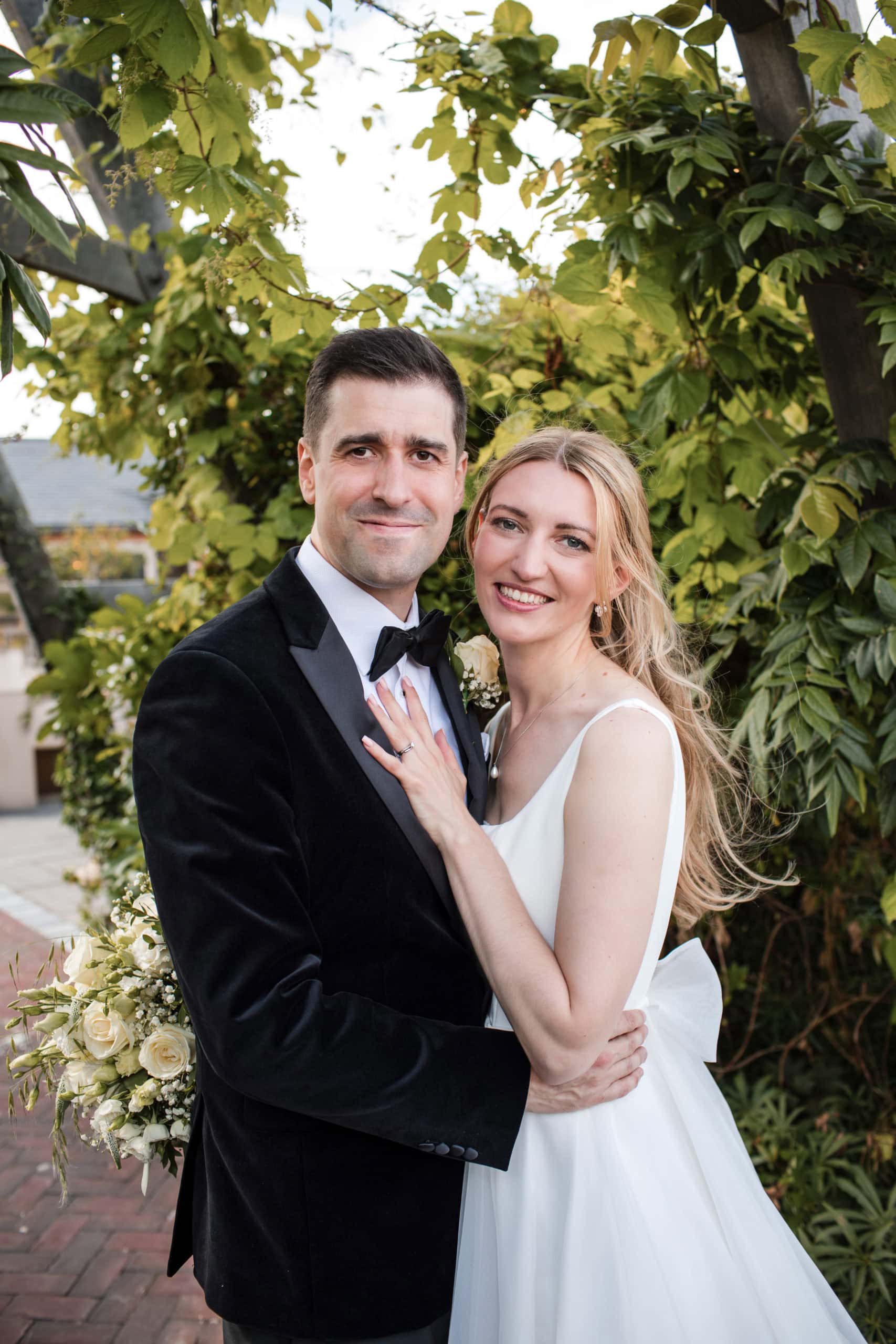 Newly married couple look to camera smiling, standing in front of green vines at Cider Mill Barns