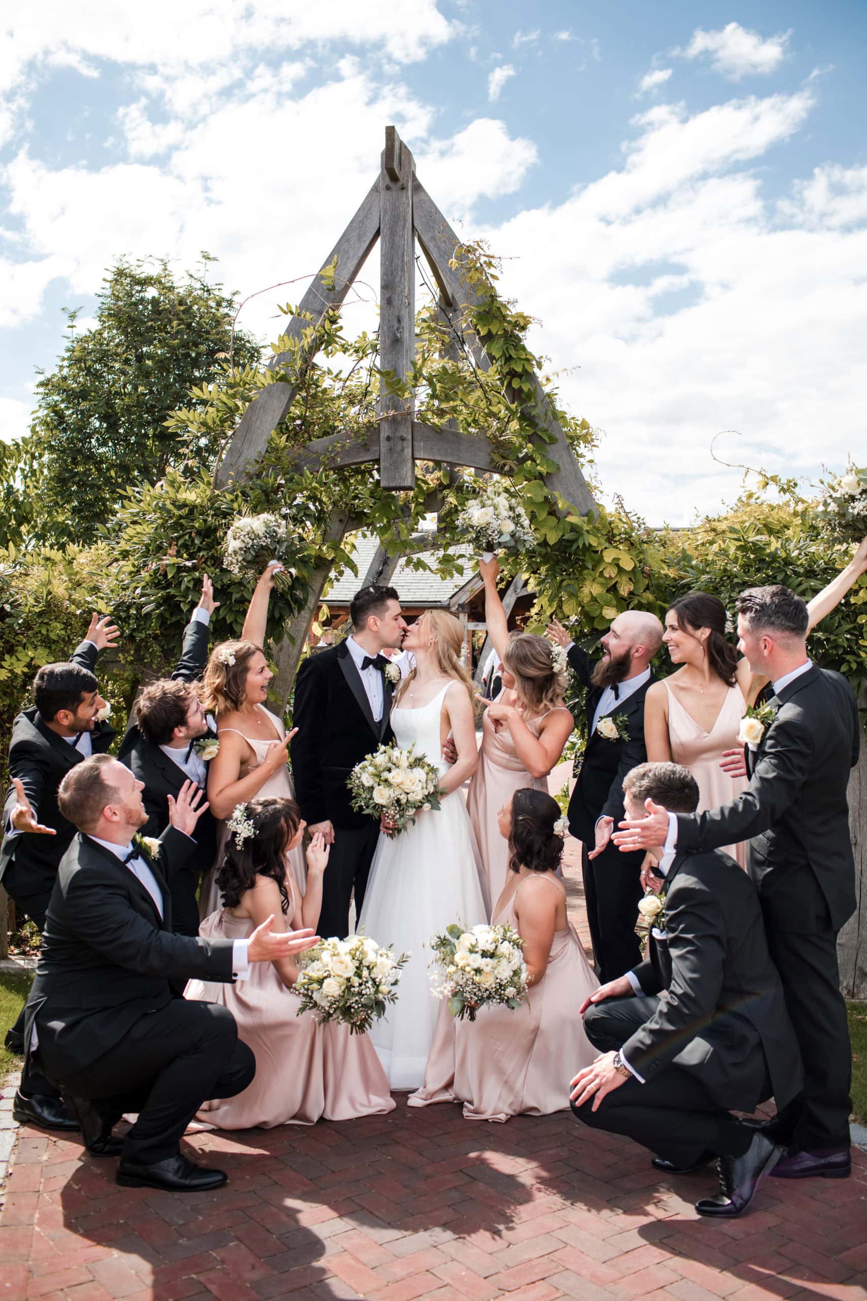 Bride and groom kiss, surrounded by their wedding party, who celebrate and cheer