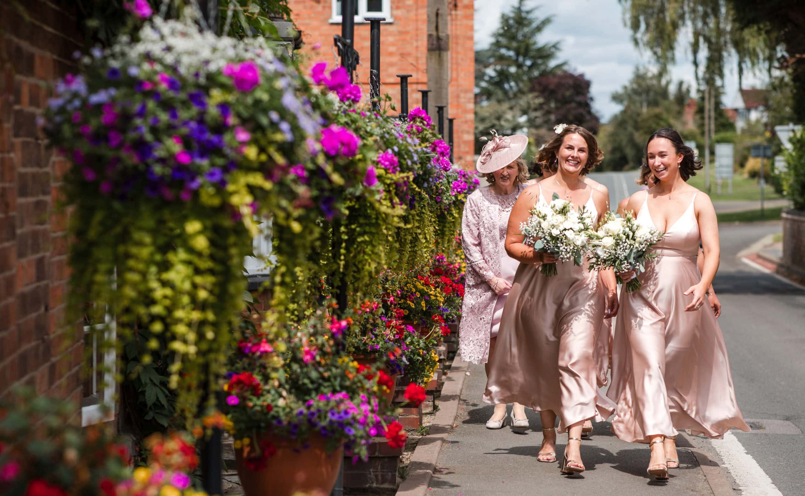 Two Bridesmaids wearing blush pink satin dresses, walk past colourful hanging flowers, in Welford on Avon, on their way to a church wedding