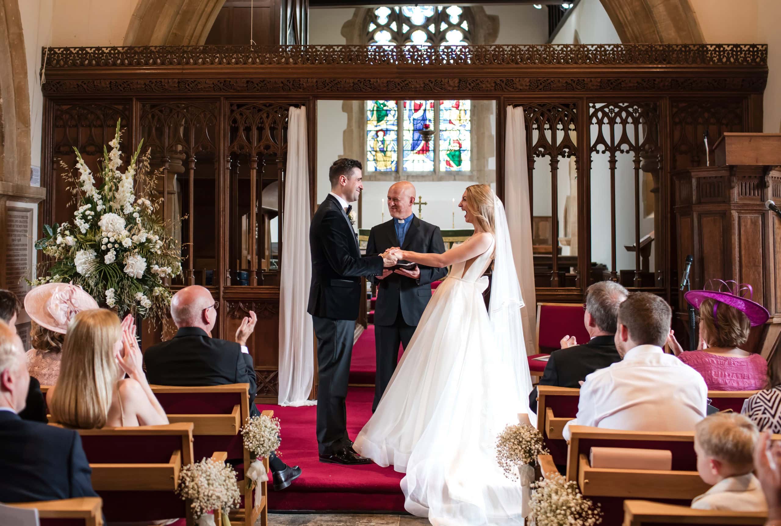 Happy Bride and Groom wearing black tuxedo stand at the alter at St Peter's Church, Welford