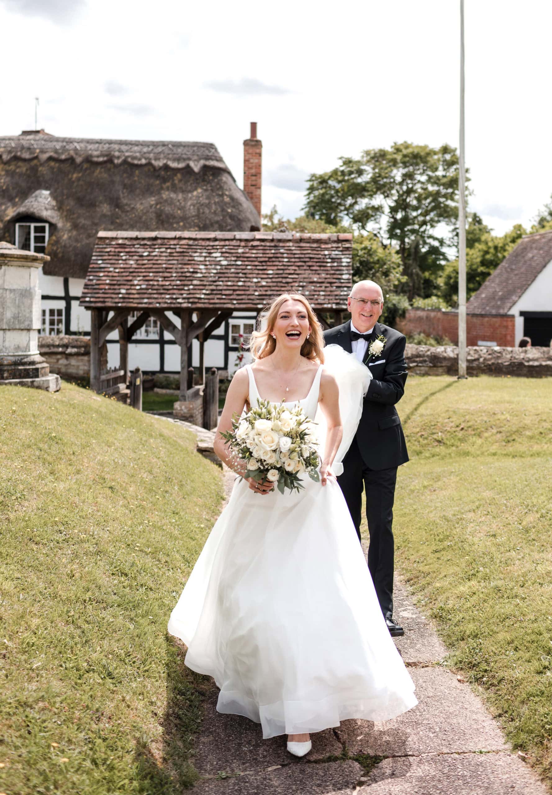 A Happy Bride walks up a path at St Peter's Church, Welford on Avon. Her Dad holds her veil and walks behind