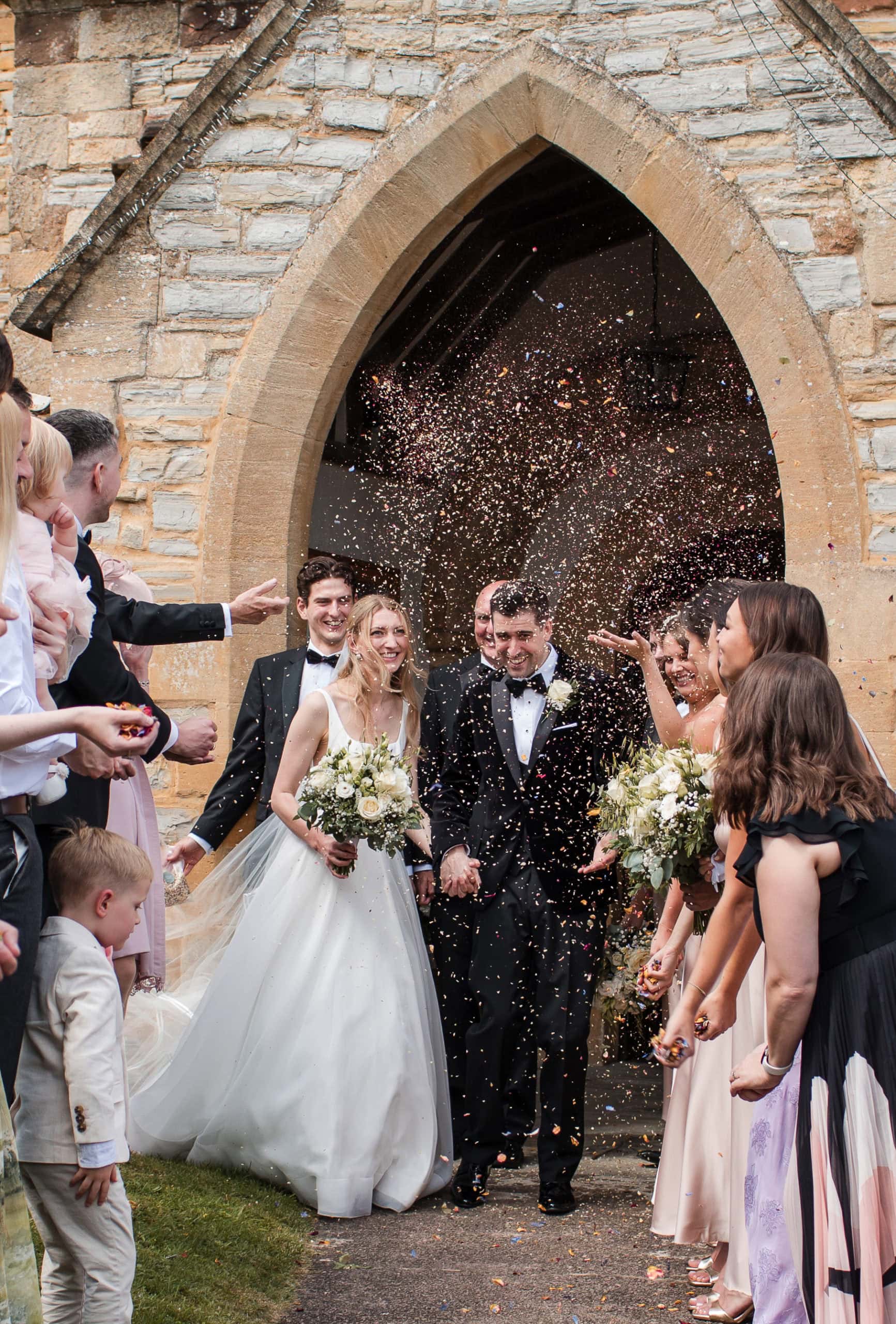 Groom wearing black velvet tuxedo holds hand with his Bride and exists St Peter's Church in Welford to lots of confetti