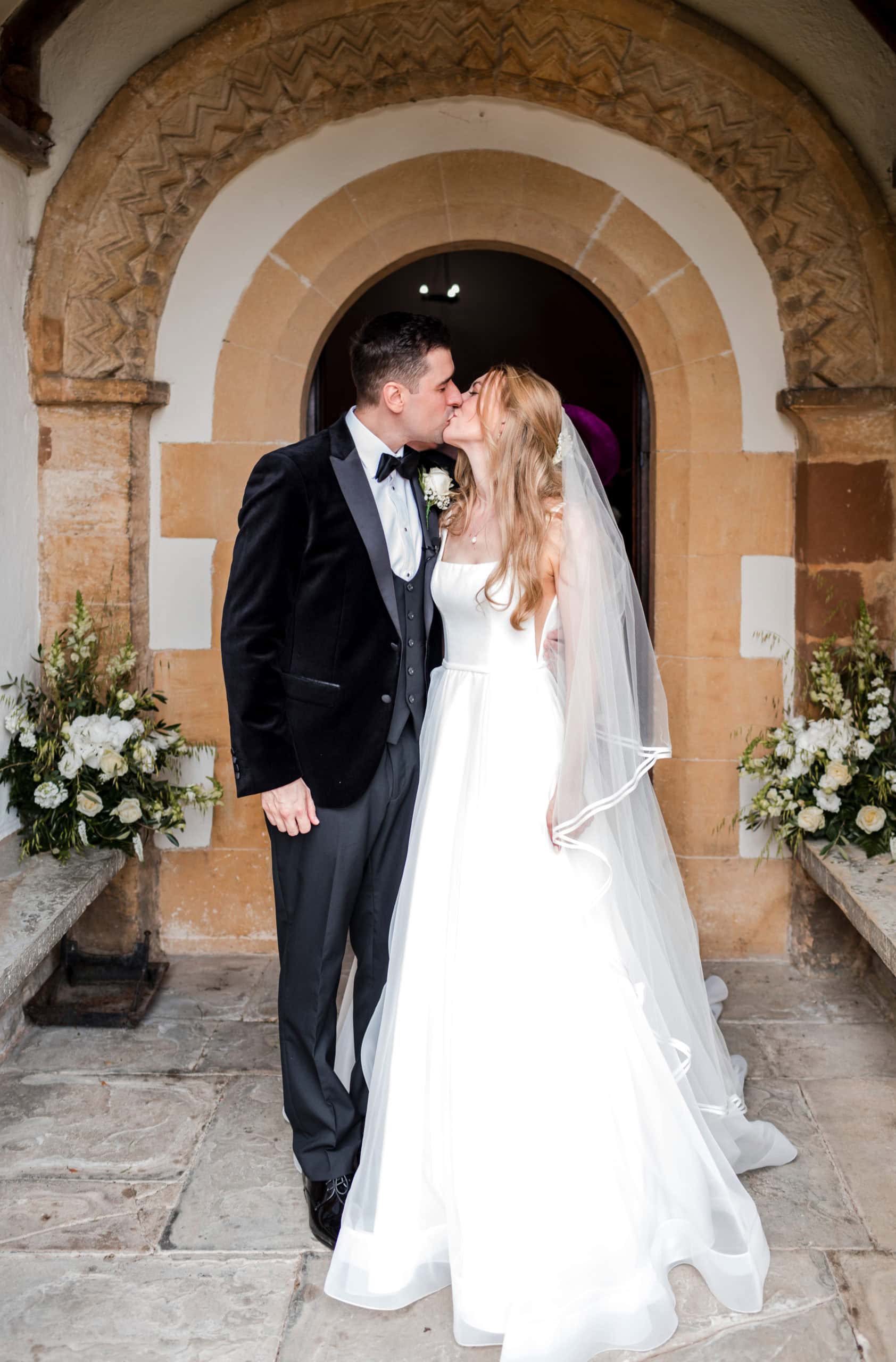 Groom wearing black velvet dinner jacket and bowtie, kisses his Bride in front of the doors of St Peter's Church, in Welford on Avon, 