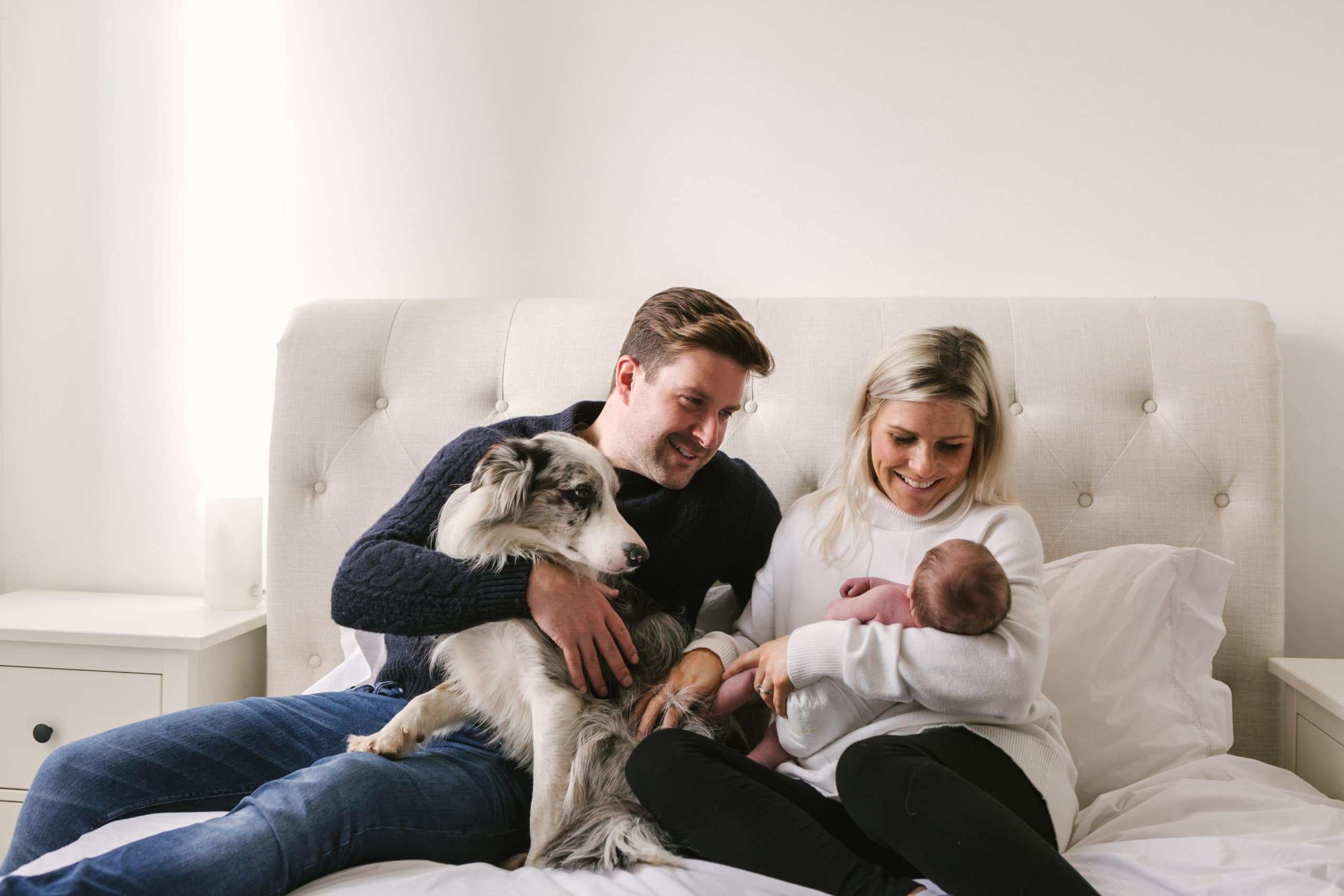 Two parents and a dog, sit on a bed to look at a newborn baby being cradled by the Mum