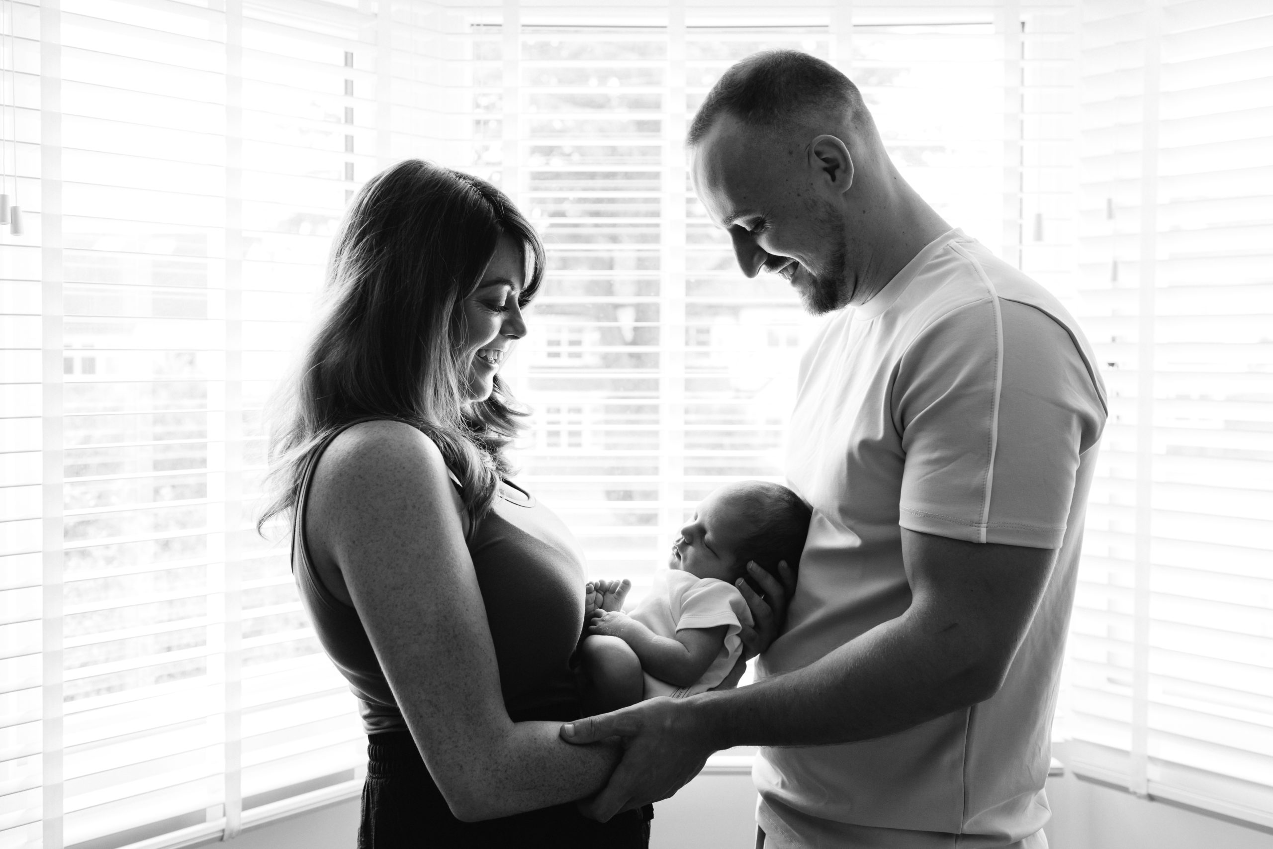 New Mum and Dad hold their newborn baby between them in front of a window