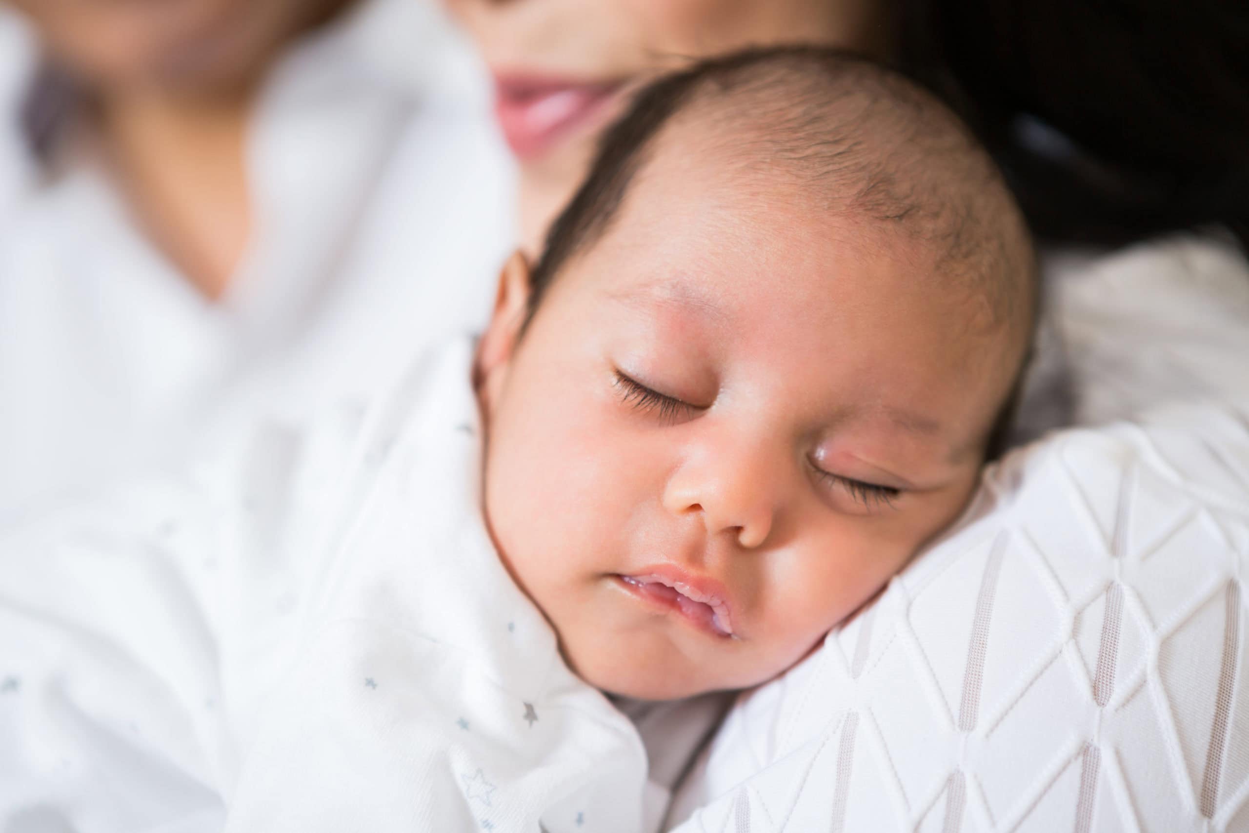 A newborn baby wearing a white babygro, asleep on his Mothers shoulder