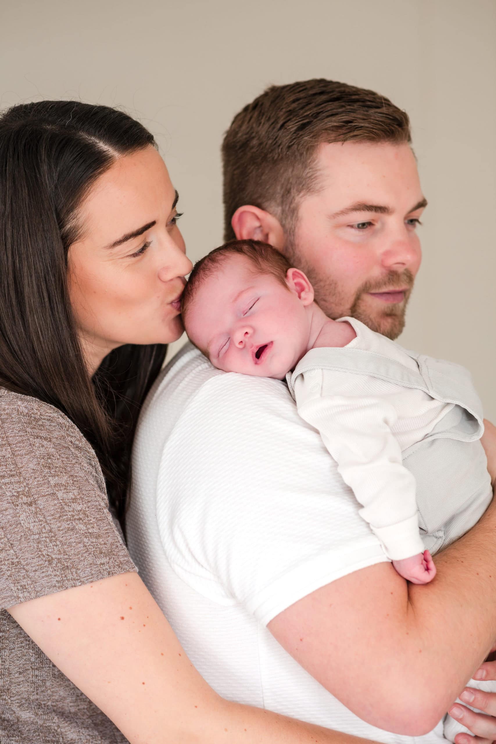 Mum wearing brown dress kisses the top of her newborn baby's head, whilst Dad wearing white T shirt holds baby on his shoulder