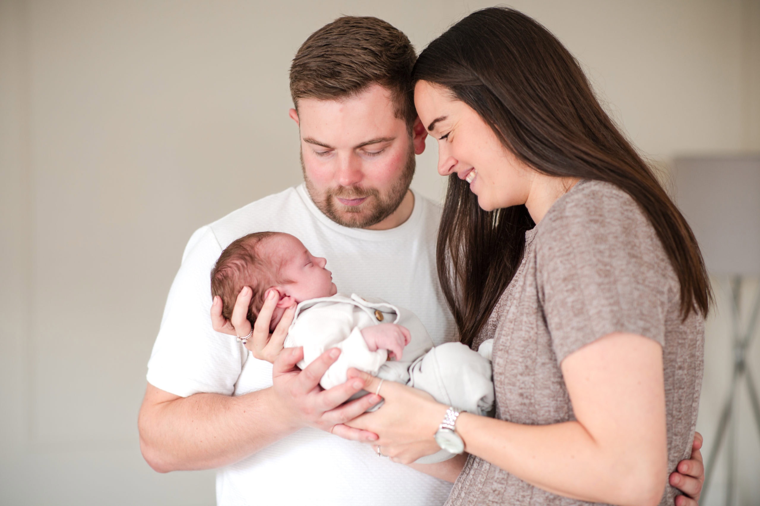 New Mum and Dad cradle and look down at their newborn baby. Both wear neutral colours