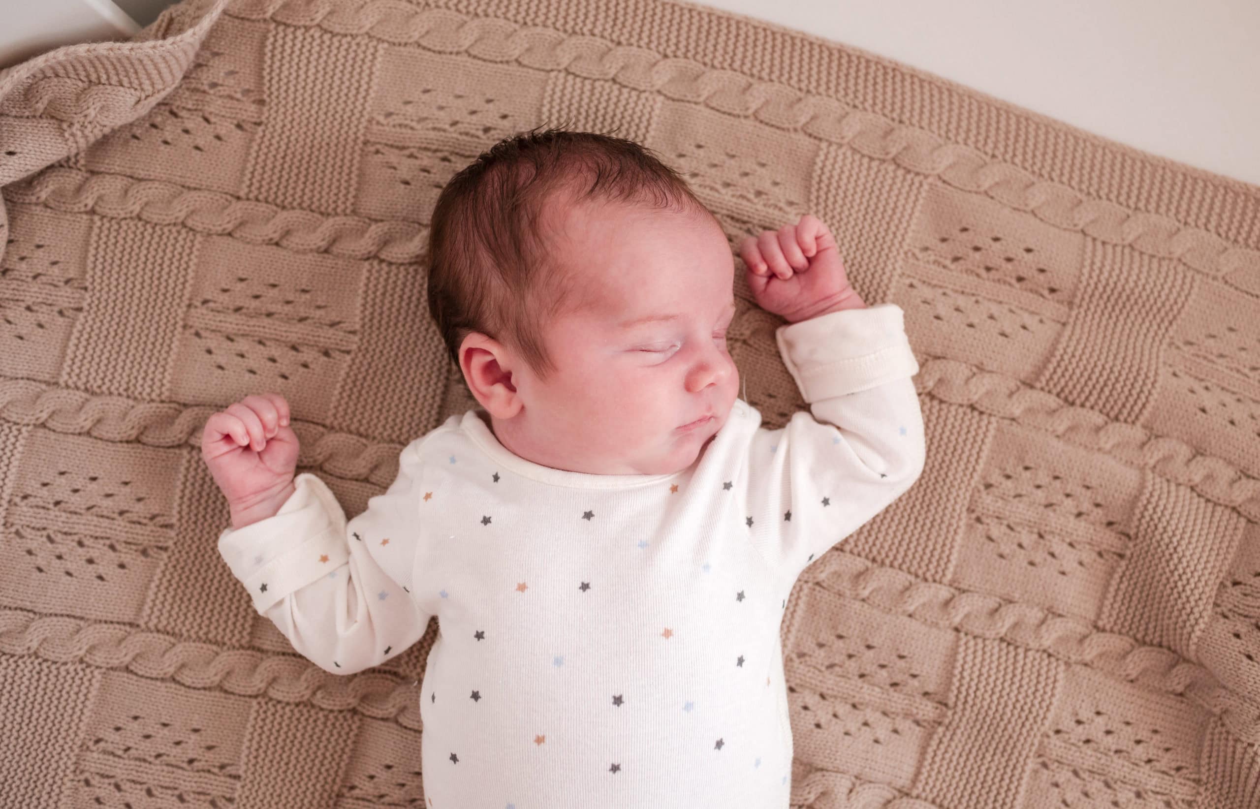 Newborn baby at home lying on a beige blanket, wearing a white babygro