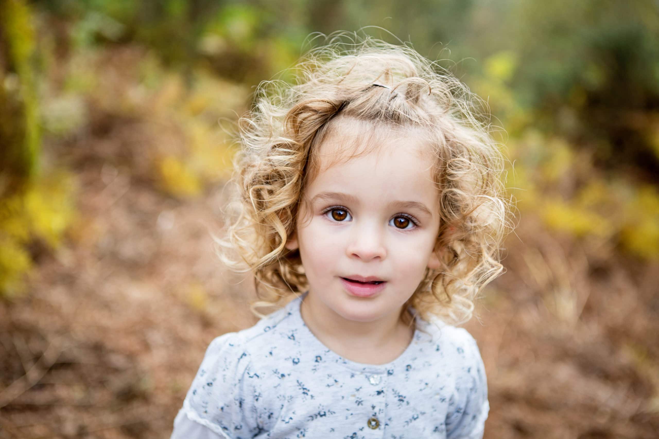 Young girl under five years old, with blonde curly hair, looks to camera in a colourful woodland backdrop