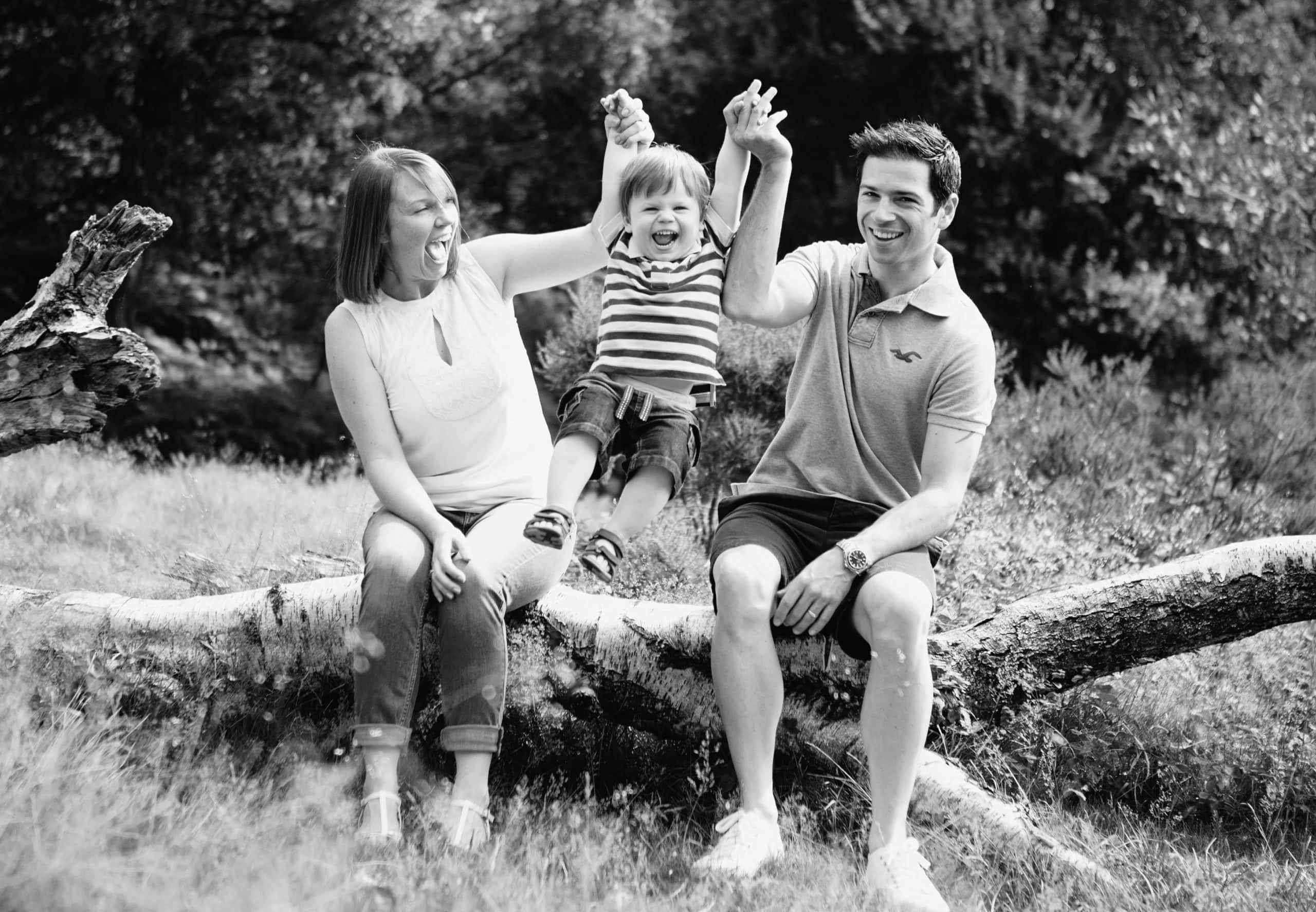 young Mum and Dad sit on a large log and lift their little boy into the air during a family photoshoot in Birmingham