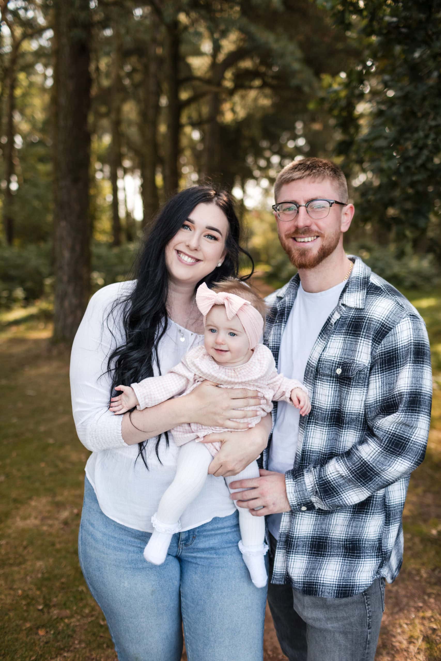 Young Dad wearing a check shirt and Mum wearing a white top, smiling, hold their six month old baby girl. Standing in woods at Lickey Hills in Birmingham