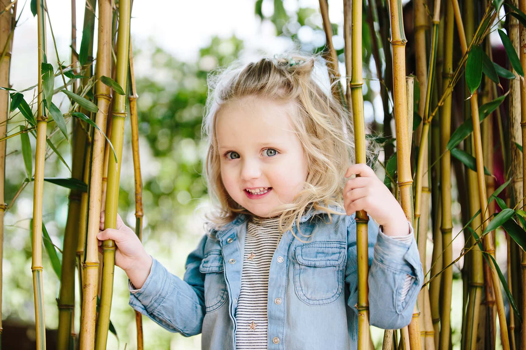 A blonde haired young girl peeps through bamboo canes at Birmingham Botanical Gardens. The girl wears a light blue denim shirt. Captured by a Birmingham family photographer