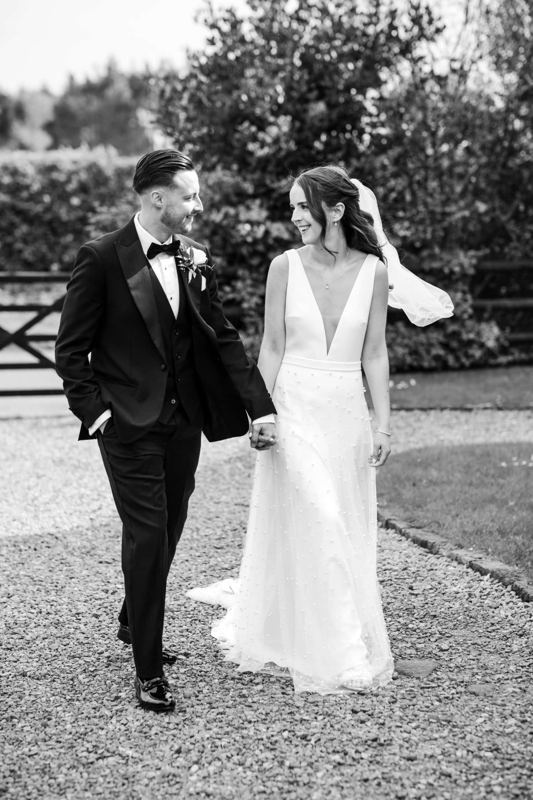 A bride and groom walking hand in hand on a gravel path, looking at each other. The bride wears a white dress with pearl details, and the groom is in a black tuxedo. Captured by Birmingham wedding photographer Jo Hastings